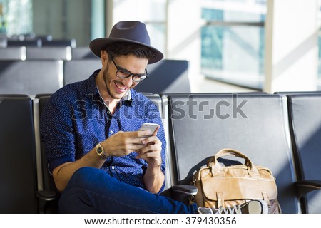 Young traveler on an airport, with a leather jacket laughing with his smartphone