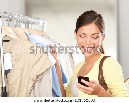 Modern woman shopping looking at smartphone texting or talking smiling happy in clothes store. Beautiful young mixed race Asian / Caucasian young woman shopper.