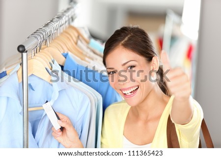 Happy shopping woman showing thumbs up while holding price tag on clothes on clothing rack. Beautiful joyful smiling multiethnic Asian Chinese / Caucasian young female shopper.
