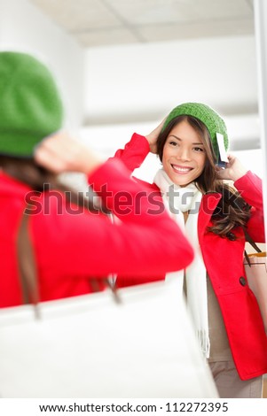 Fall / Winter shopping woman trying on knit hat looking in mirror smiling happy holding shopping bags inside in clothing store. Multiracial shopper woman.