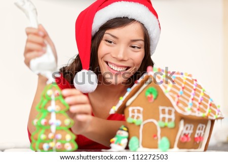Girl making Christmas gingerbread house. Young woman in Christmas preparations putting icing on gingerbread house. Model wearing santa hat.