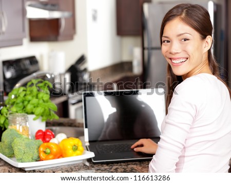 Woman Using Laptop Computer In Kitchen With Greens And Vegetables. Lifestyle Cooking Concept With Smiling Happy Mixed-Race Asian Chinese Caucasian Woman Working Sitting In Her Kitchen.