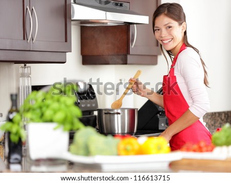 Cooking woman in kitchen stirring in pot making food for dinner. Young housewife smiling happy looking at camera. Mixed-race Caucasian / Asian chinese woman in her twenties.