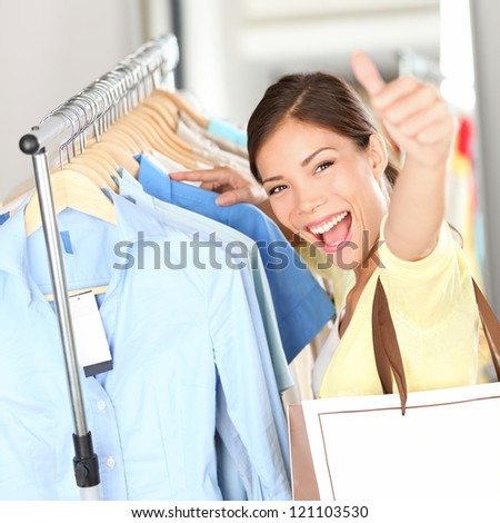 Shopping - Happy shopper woman showing thumbs up excited holding shopping bag in clothing store looking for clothes on sale. Beautiful mixed race asian caucasian woman model.