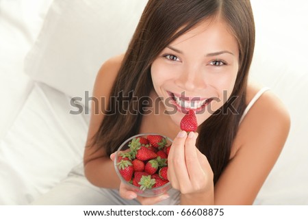 Portrait of woman eating strawberries. Healthy happy smiling woman eating strawberry inside in bed holding a bowl of strawberries. Gorgeous smile on mixed Caucasian Asian female model.
