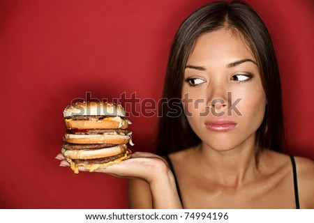 Unhealthy eating - junk food concept. Woman looking at big oversized burger thinking. Pretty Caucasian Asian model over red background.