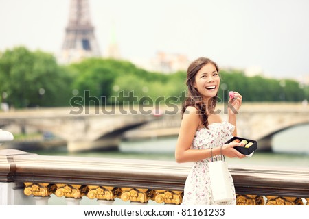 Paris woman smiling eating the french pastry macaron in Paris. Eiffel tower and Pont Des Invalides in the background. Cute beautiful mixed race Asian Caucasian female model.