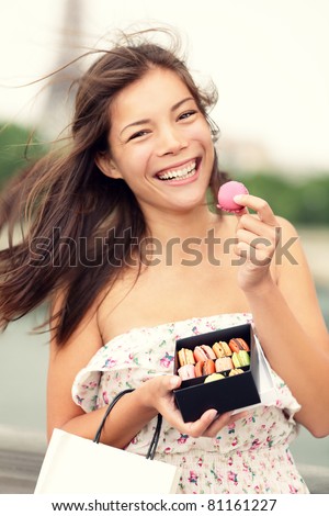 Paris woman eating macarons in Paris happy and smiling. Eiffel tower in the background. Cute beautiful mixed race Asian Caucasian female model playful in dress summer dress holding small shopping bag.