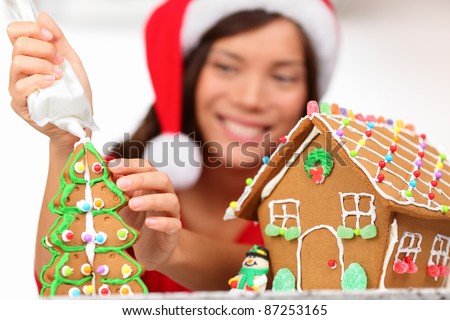 girl making gingerbread house. Young woman in Christmas preparations putting icing on gingerbread house. Model wearing santa hat.