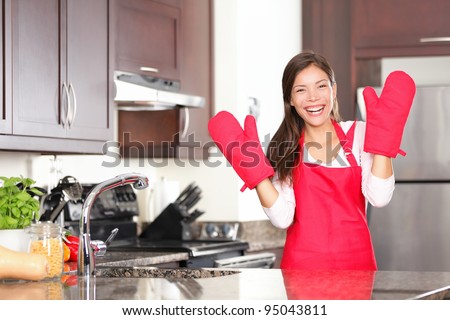 Happy baking cooking woman standing in her new kitchen smiling cheerful wearing apron and oven mitts ready to bake. Beautiful young mixed race Caucasian / Chinese woman at home.