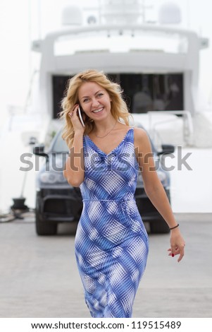 Wealthy young woman walks talking on the phone with expensive car and yacht on the background.