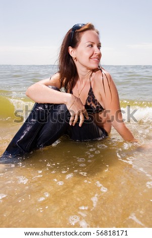 Young adult woman in wet jeans sits in splashes of tidal waves of a sea smiling