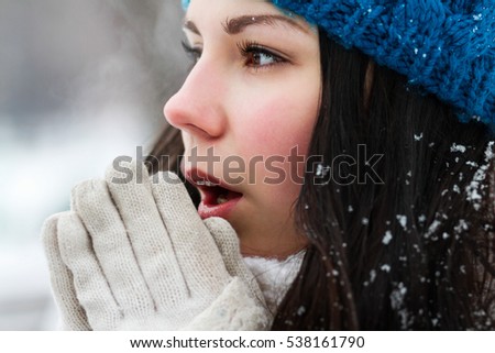Portrait of frozen white brunette girl in knit blue hat.Young woman breathe with warm air on her frozen hands.Close up.Pretty female model outside in cold winter day.Cute European chick freezing