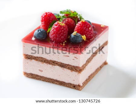 Slice of delicious raspberry mousse cake made from layers of chocolate genoise, glazed with raspberry jelly, and decorated with blueberries, raspberries, sprig of chervil, and pieces of gold leaf