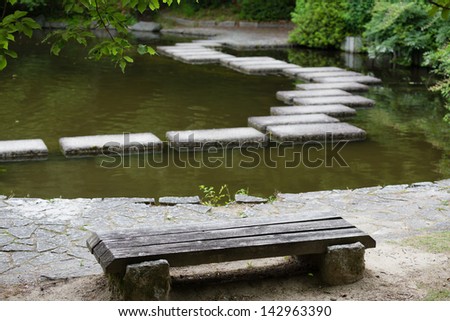 Zen stone path in a Japanese Garden across a tranquil pond with a stone bench on foreground