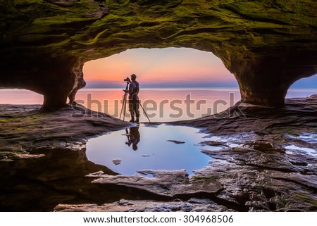 A photographer with tripod, standing in the mouth of a Michigan sea cave, is silhouetted by a colorful sunset over Lake Superior.