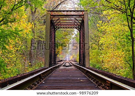 Railroad tracks lead through an iron trestle (bridge) toward infinity with changing fall leaves surrounding.