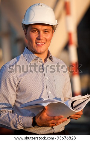portrait of well-dressed man with documants in hard hat standing against the construction