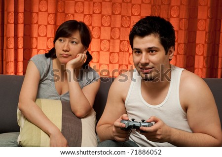 young couple sitting on a couch. The guy is playing video games, while the girl is bored