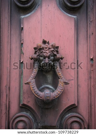 Old doorknocker with the garlanded head of a man or demon mounted on an old wooden door with a light coating of pink paint