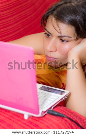 Woman lying on her stomach on a red sofa concentrating as she uses a pink laptop