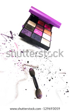 Artistic arrangement of colourful eye makeup in a box with an applicator and scattered remnants and squiggles of powdered cosmetic in the foreground on white