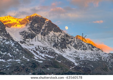 view from St. Moritz in Switzerland: high snow and ice capped alpine mountain range with red illuminated peaks at sunset and the moon out