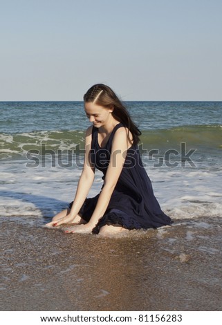 Beautiful young girl in black wet dress sits in water of the sea