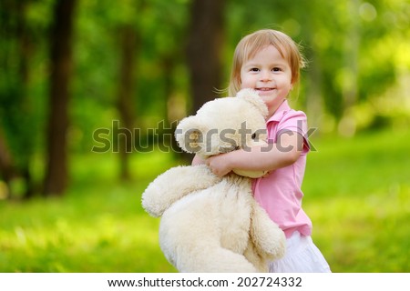 Adorable little toddler girl with a big teddy bear having fun in summer park on be?utiful sunny day