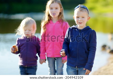 Two little sisters and their brother eating buns, laughing and joking by a river at sunny autumn day
