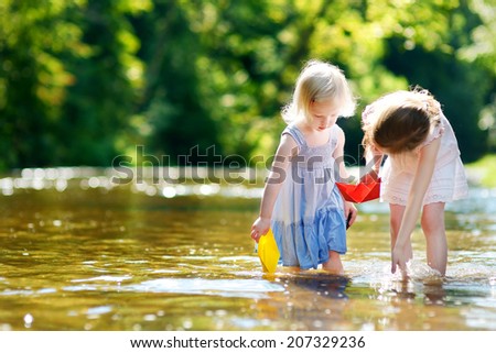 Two adorable little sisters playing with paper boats by a river on warm and sunny summer day