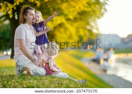 Young father and his two little daughters sitting by a river and watching canoes passing by