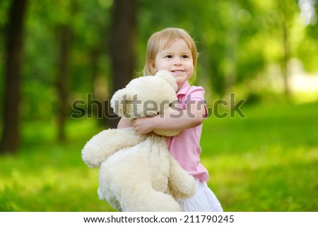 Adorable little toddler girl with a big teddy bear having fun in summer park on beautiful sunny day