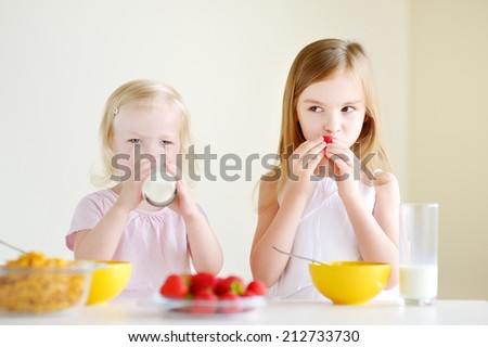 Two cute little sisters eating cereal with strawberries and drinking milk in white kitchen