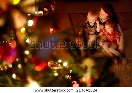 Young mother and her two little daughters sitting by a Christmas tree in a cozy dark living room on Christmas eve