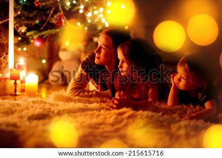 Young mother and her two little daughters sitting by a fireplace in a cozy dark living room on Christmas eve