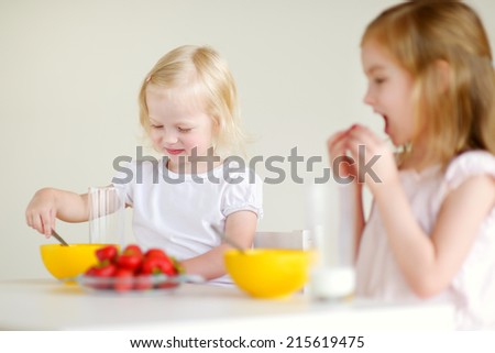 Two cute little sisters eating cereal with strawberries and drinking milk in white kitchen
