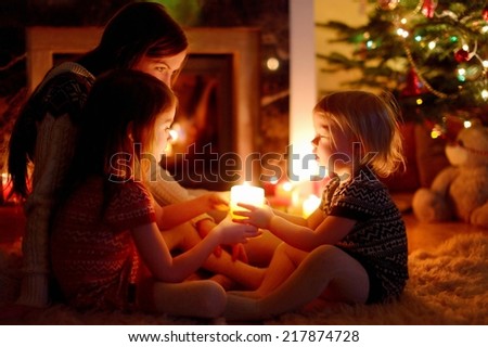 Young mother and her two little daughters sitting by a fireplace holding candles in a cozy dark living room on Christmas eve