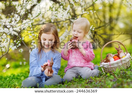 Two adorable little sisters eating chocolate bunnies in a spring garden on Easter day