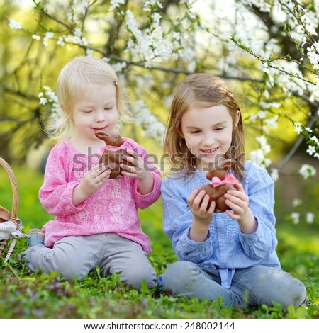 Two adorable little sisters eating chocolate bunnies in a spring garden on Easter day