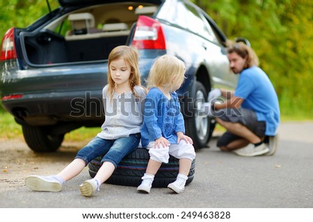 Two adorable little sisters sitting on a tire and waiting while their father is changing a car wheel outdoors on beautiful summer day