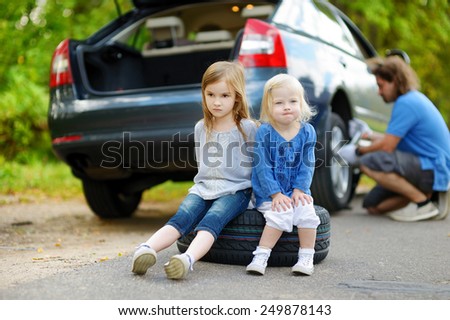 Two adorable little sisters sitting on a tire and waiting while their father is changing a car wheel outdoors on beautiful summer day