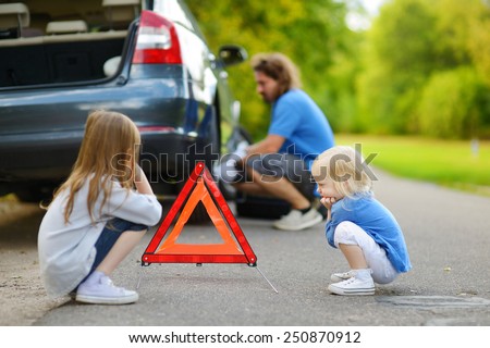 Adorable little girls waiting by the red warning triangle sign while their father is changing a car wheel outdoors on beautiful summer day