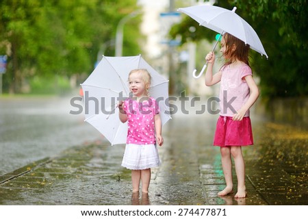 Two cute little sisters standing in a puddle holding umbrella on a rainy summer day