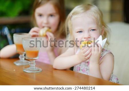 Two little sisters drinking orange juice and eating pastries in an outdoor cafe on warm and sunny summer day