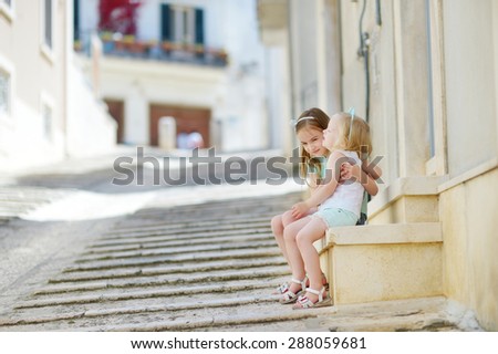 Two adorable little sisters laughing and hugging on warm and sunny summer day in italian town