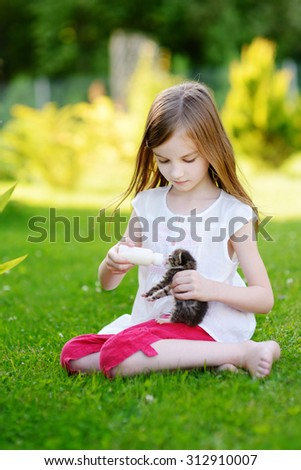 Adorable little girl feeding small kitten with kitten milk from the bottle at summer day