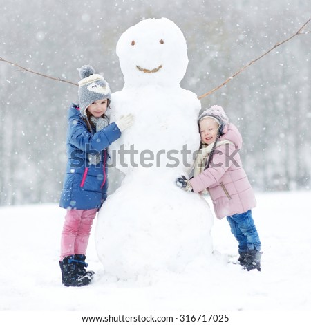 Two funny adorable little sisters building a snowman together in beautiful winter park during snowfall
