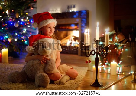 Happy little girl sitting by a fireplace in a cozy dark living room on Christmas eve