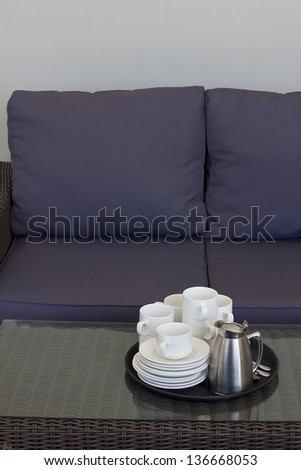 A tray with empty coffee cups and a full coffee pot and sugar bowl placed on a rattan table with a glass top, in front of a rattan sofa with blue cushions